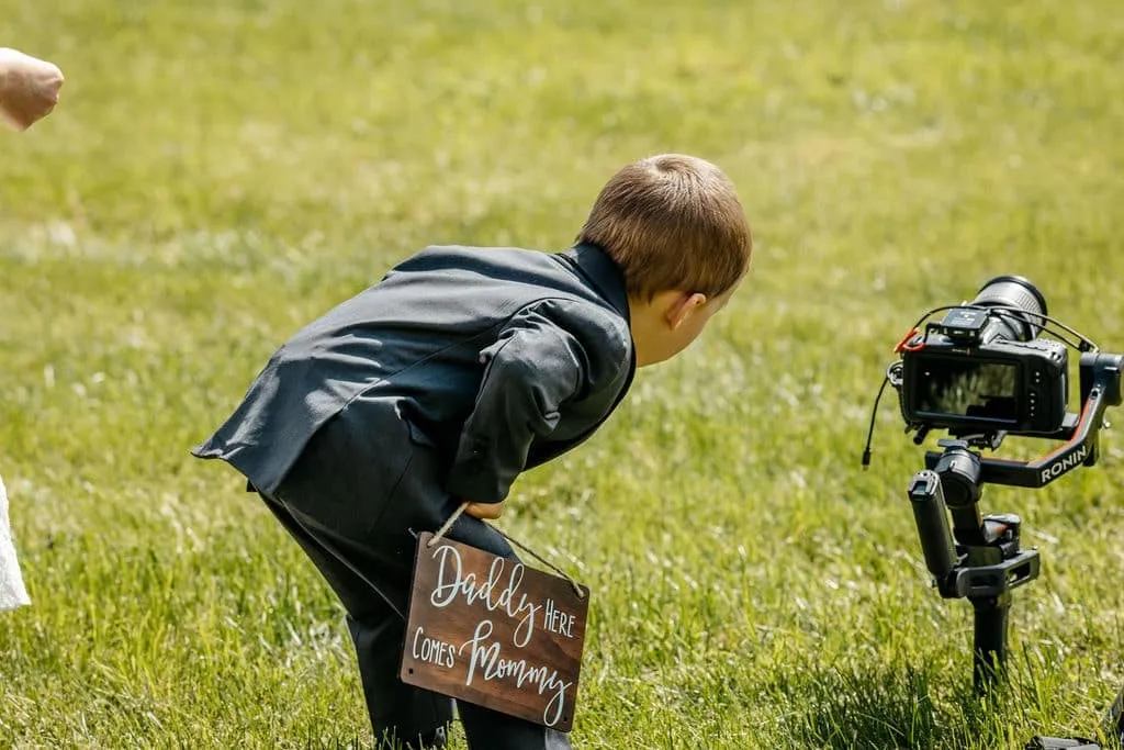 Ring bearer with "Daddy here comes Mommy" sign filmed by the best wedding photographer in New England during outdoor ceremony | Love in Focus Studio