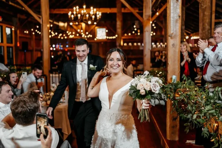 Smiling bride and groom enter a rustic barn-style wedding reception in New England, surrounded by guests and string lights | Love in Focus Studio