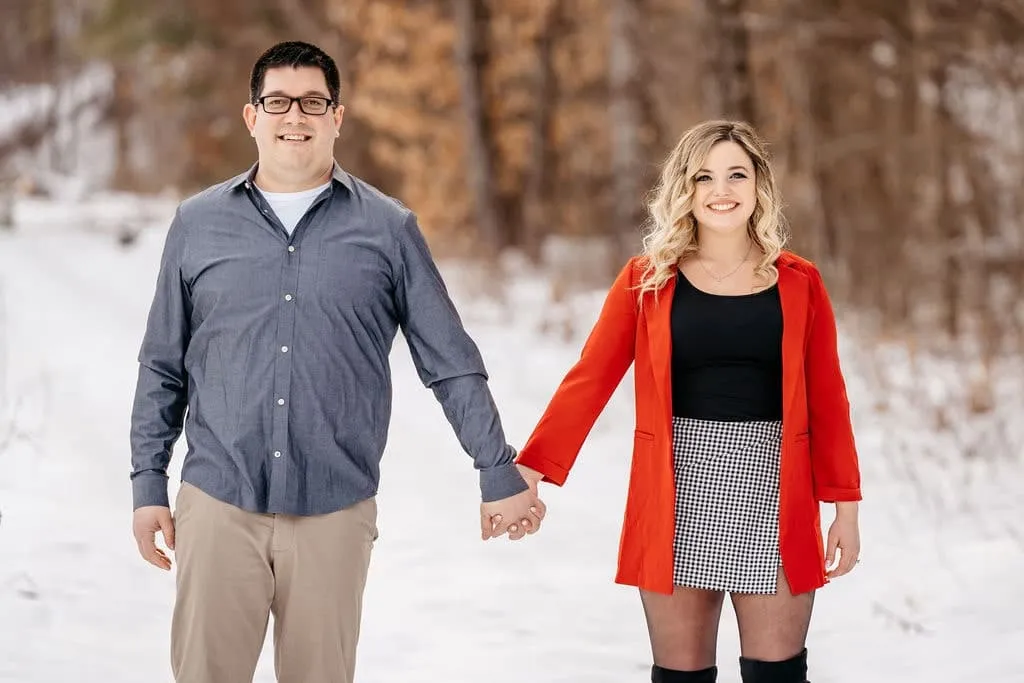 Smiling couple holding hands during a winter engagement photoshoot; woman in red coat and checkered skirt, man in blue shirt and khakis, snowy forest background | Love in Focus Studio