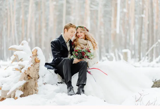 Winter bride with a flower crown and burgundy bouquet beside her groom in a snowy forest
