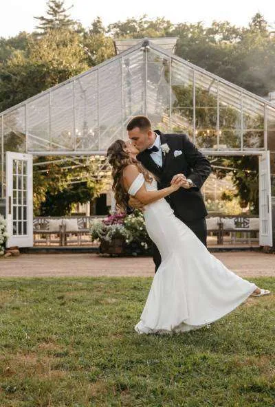A groom dips and kisses his bride on a lawn in front of a beautiful, classic glass greenhouse structure during a New England wedding celebration.