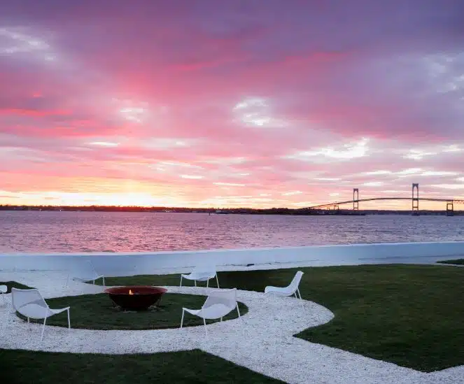A fire pit on the lawn at Belle Mer in Newport, Rhode Island, with a pink and purple sunset over the water and bridge.