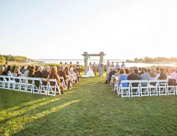 An outdoor wedding ceremony at Harbor Lights in Warwick, Rhode Island, with guests seated on a lawn overlooking the water at sunset.