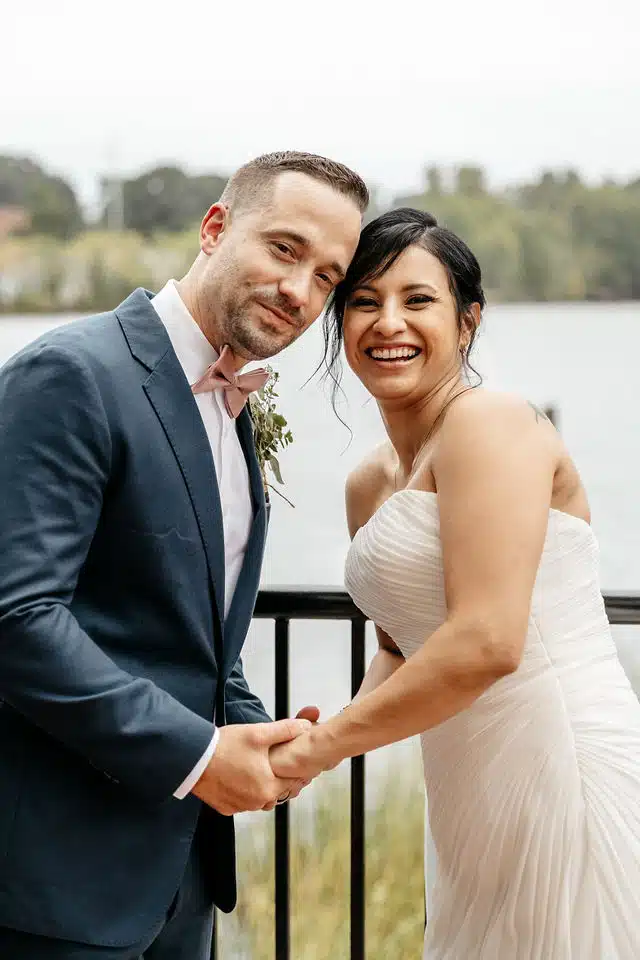 A smiling bride and groom hold hands at Waterman Grille Restaurant in Providence, Rhode Island, with a river in the background.