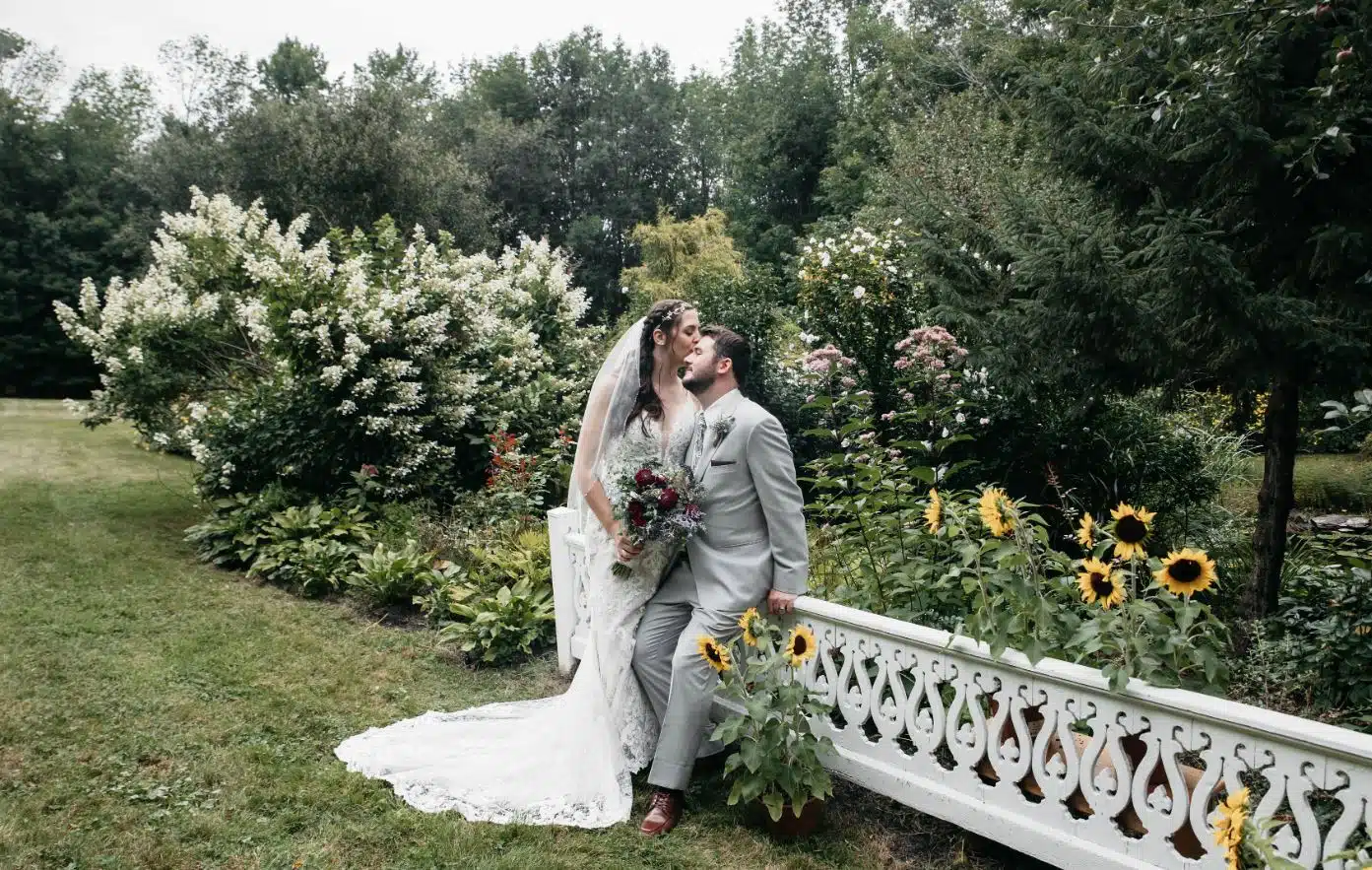 A bride and groom kiss in the garden, surrounded by sunflowers, during their wedding at The Barn at Seven Elms in Lebanon, Maine.