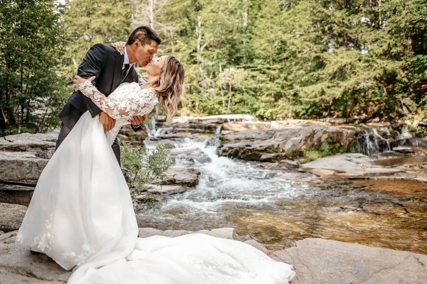 A groom dips his bride for a kiss by a waterfall during their wedding at The Barn at Whitney's Inn in Jackson, New Hampshire.