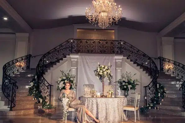 A bride sits at a sweetheart table at Aria in Prospect, CT, in front of a grand staircase and under a chandelier.