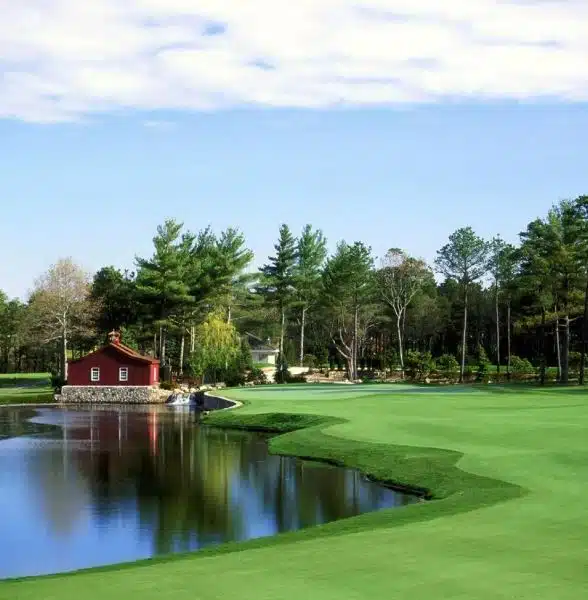 A scenic view of the golf course at Willowbend Country Club in Mashpee, MA, with a red barn next to a tranquil pond.