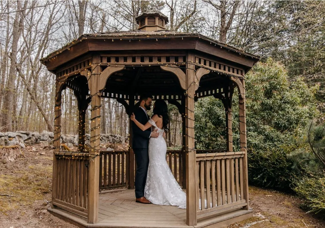 A bride and groom embrace inside a rustic wooden gazebo at Granite Rose by Wedgewood Weddings in Hampstead, NH.
