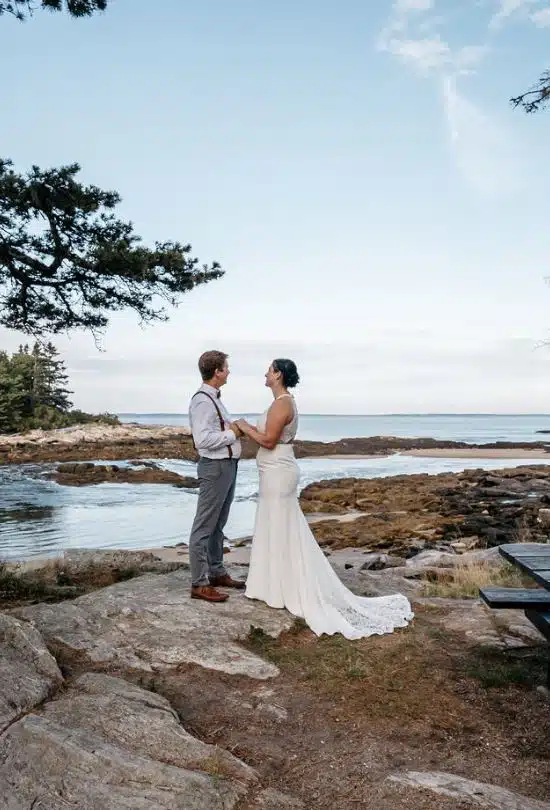 A bride and groom hold hands on the rocky coast during their wedding elopement at La Verna Preserve in Bristol, Maine.
