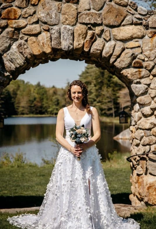 A bride stands under a stone archway, holding a bouquet, during her wedding at Lakefalls Lodge in Munsonville, New Hampshire.