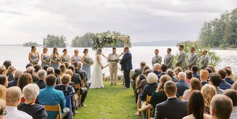 An outdoor wedding ceremony at Migis Lodge, with guests watching the couple by the shore of Sebago Lake in South Casco, Maine.