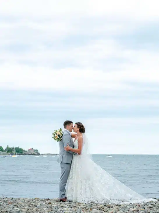 A bride and groom stand on a rocky beach at the Oceanview of Nahant in Massachusetts, with the ocean behind them.
