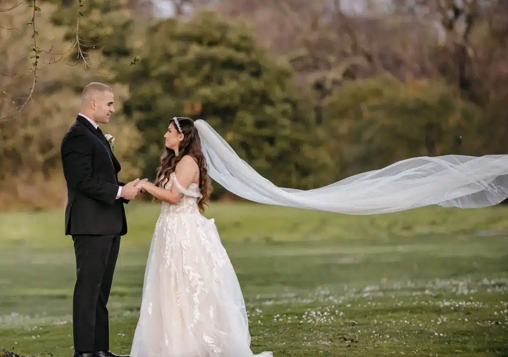 A bride and groom hold hands in a field at The Orchards Hotel in Williamstown, Massachusetts, with her long veil flying in the wind.