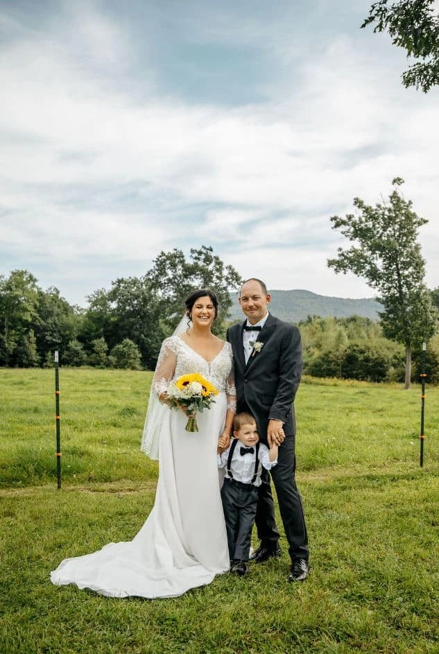 A bride and groom pose with their young son in a field during their wedding at Tumbledown Farm in Brookfield, New Hampshire.