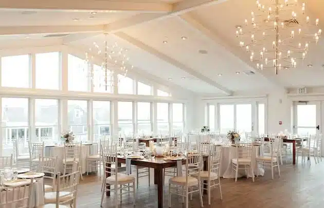 A bright, airy wedding reception room at Wychmere Beach Club in Harwich Port, MA, with large windows and modern chandeliers.