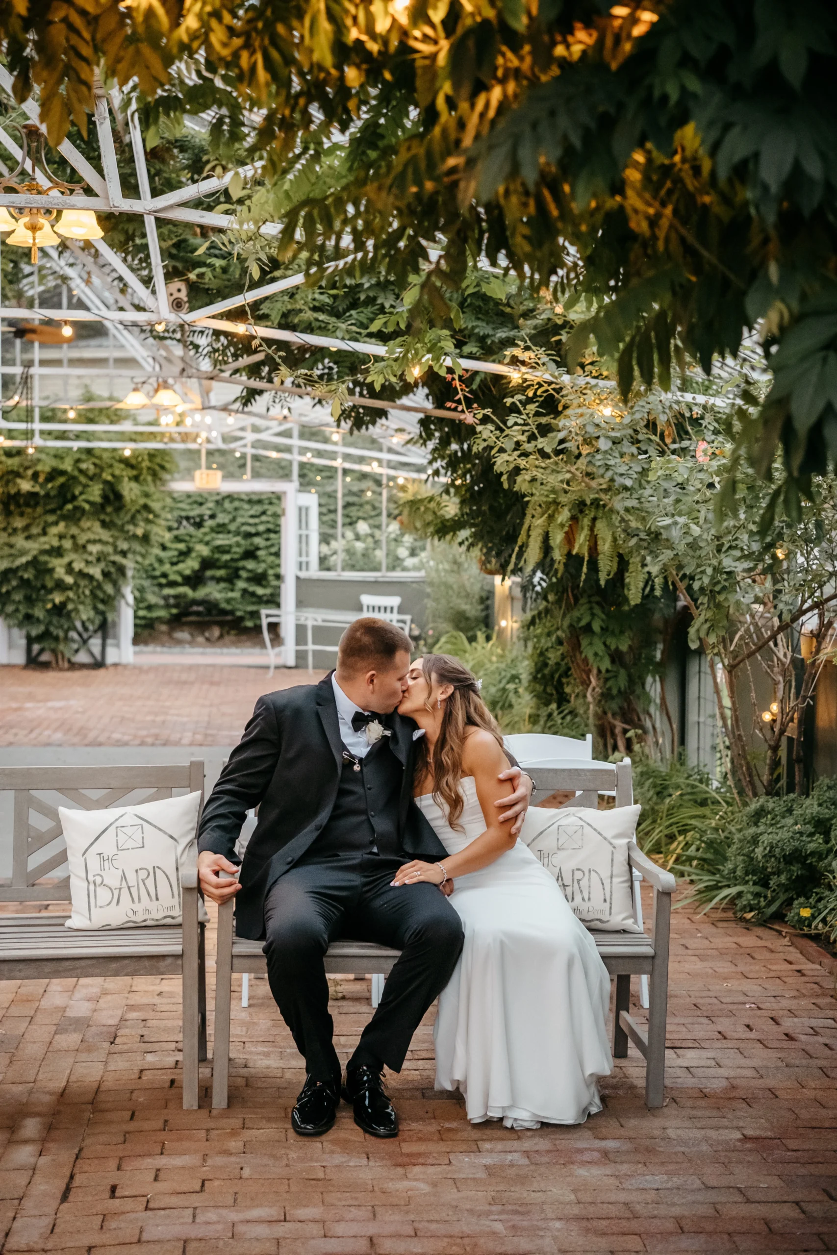 A bride and groom kiss on a bench in the greenhouse courtyard at The Barn on the Pemi in Plymouth, New Hampshire.