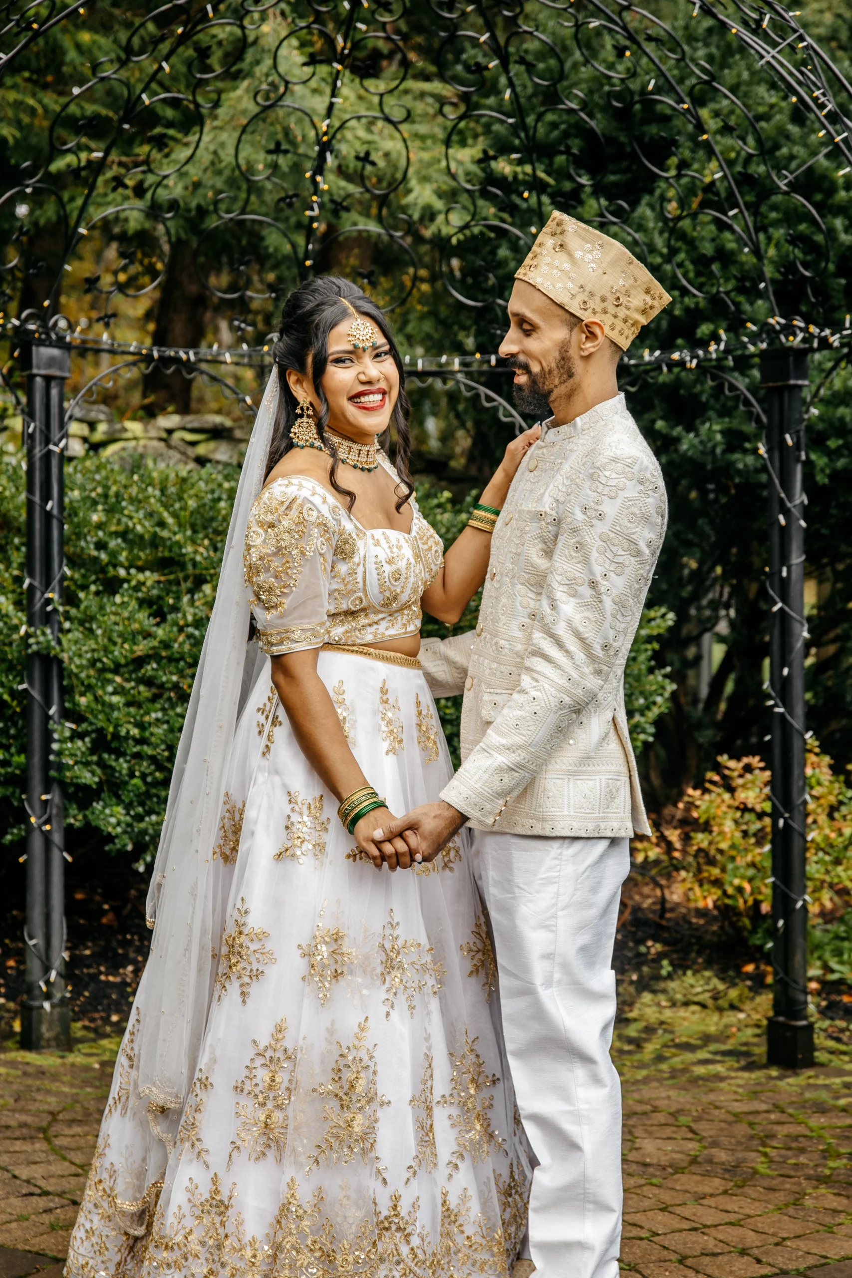 A smiling bride and groom in white and gold traditional Indian attire hold hands at Granite Rose by Wedgewood Weddings, NH.