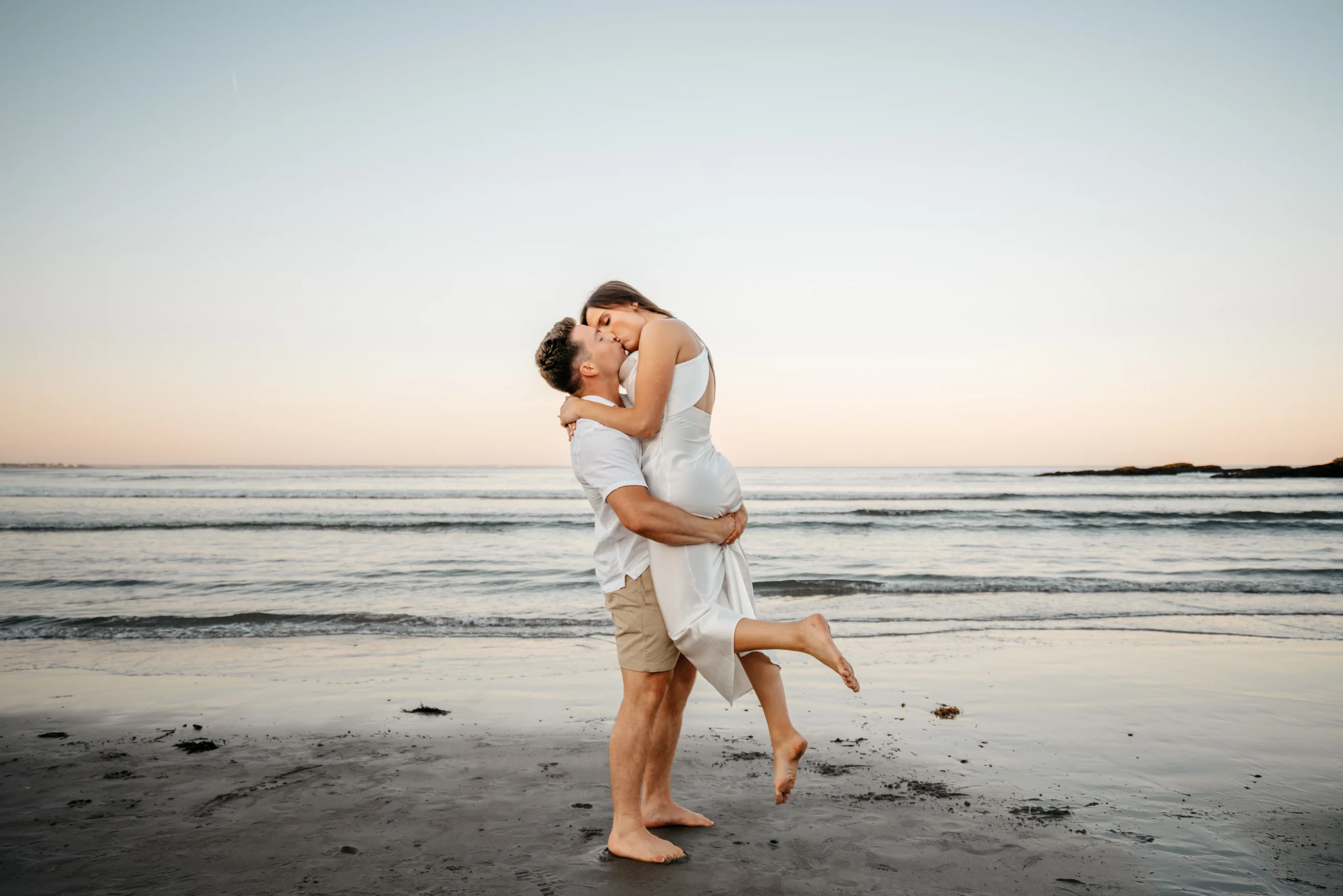 A man lifts his fiancée for a kiss on the beach at sunset during their Marginal Way engagement session in Ogunquit, Maine.