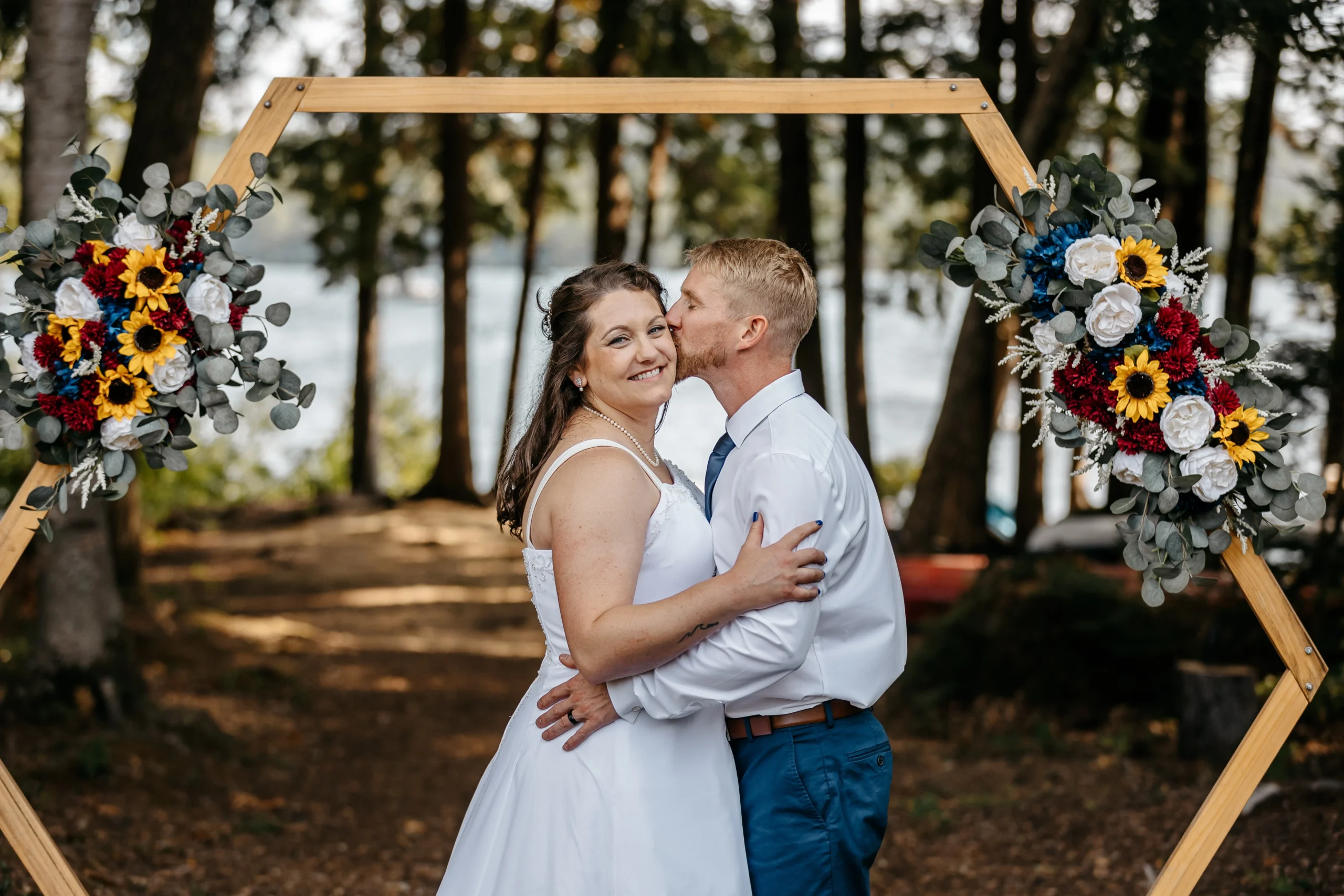 A groom kisses his bride's cheek as they stand under a hexagonal wedding arch at Moose Lake Ranch in Otisfield, Maine.