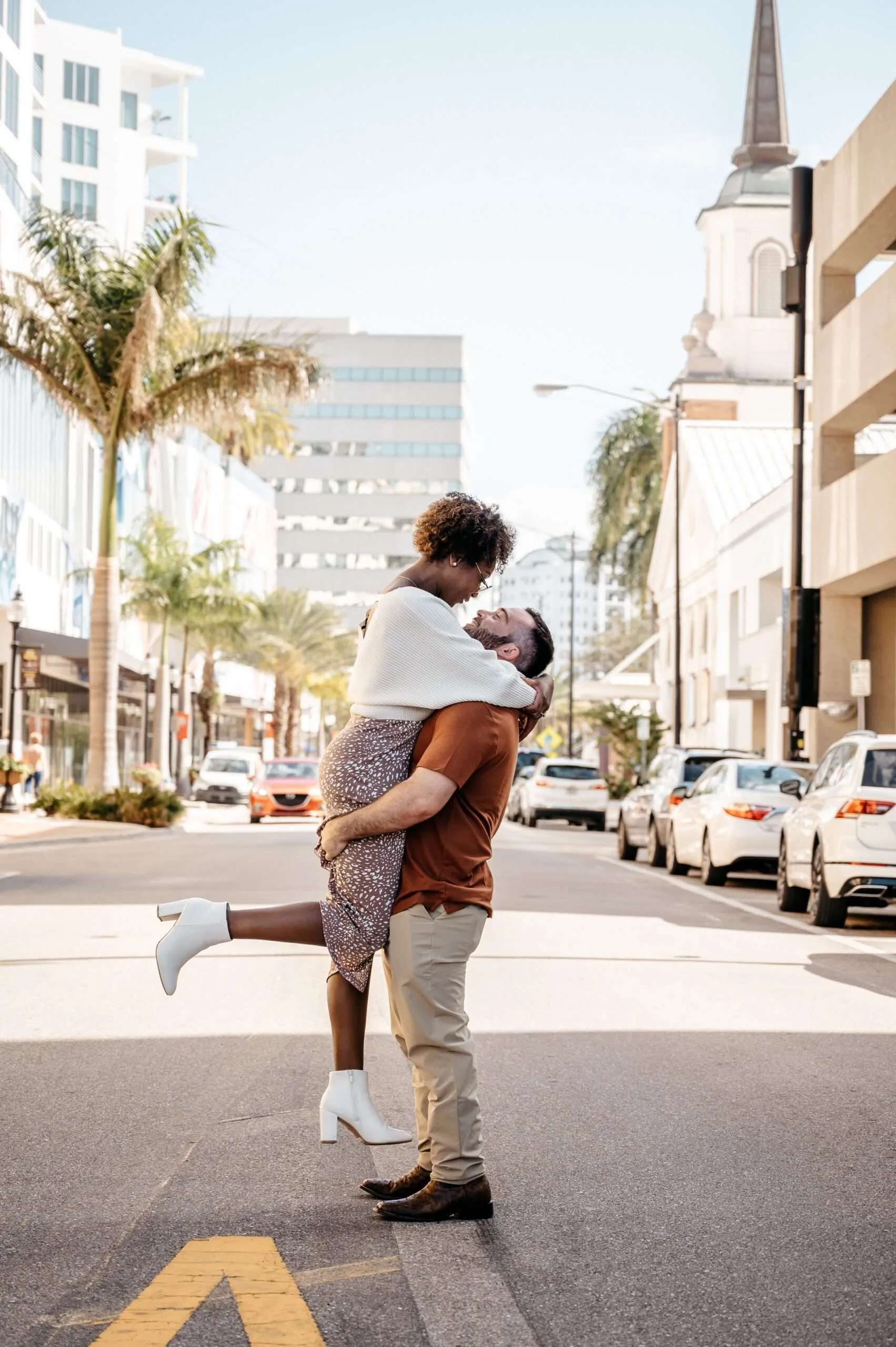Interracial couple embracing and kissing in the middle of a sunlit city street, surrounded by palm trees and urban buildings during a romantic engagement photoshoot.