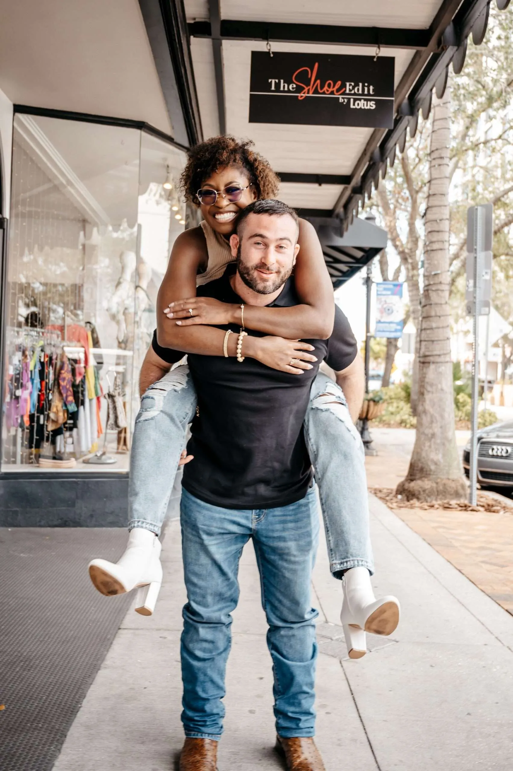 Happy interracial couple sharing a playful piggyback on a city sidewalk, smiling in front of boutique storefronts during an urban engagement photoshoot.