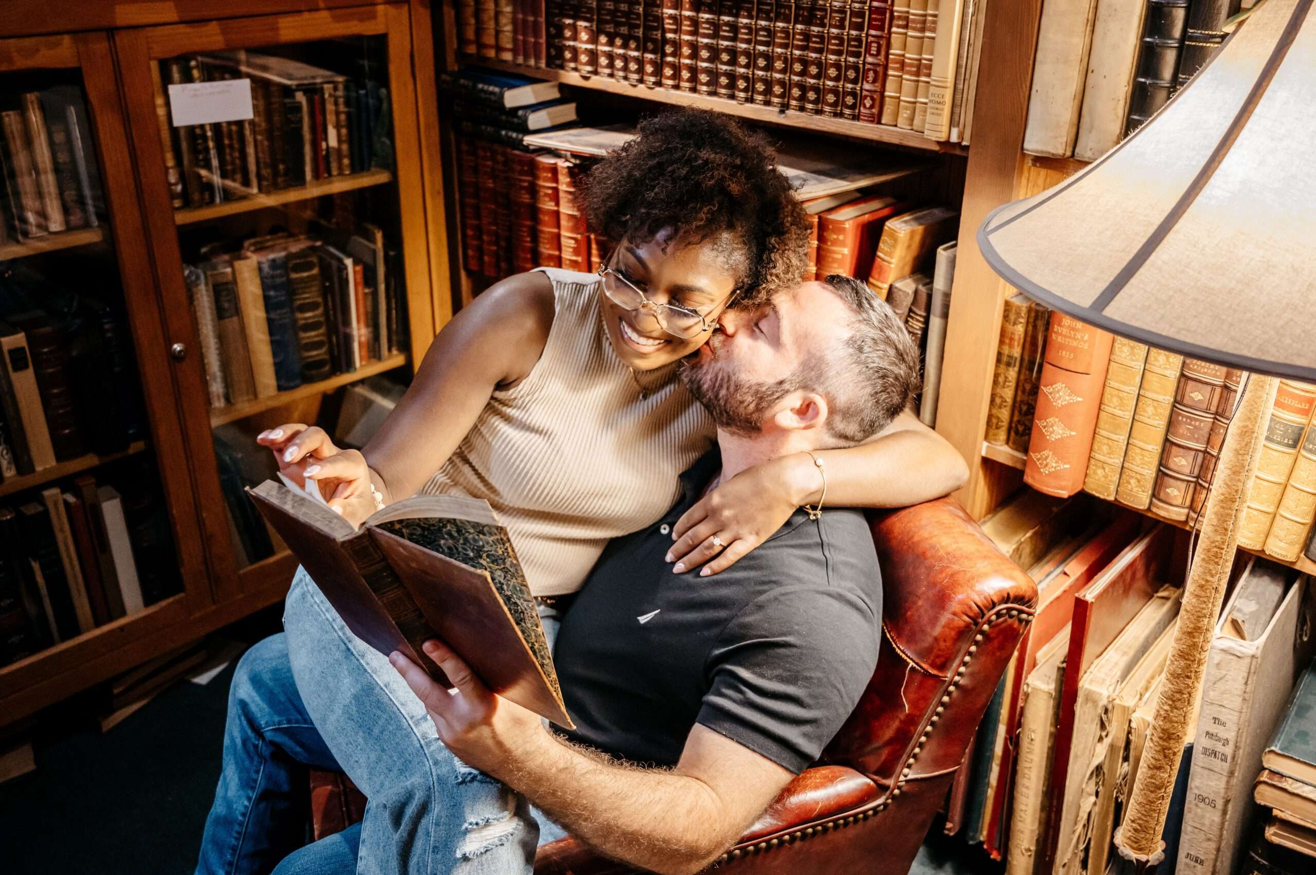 Interracial couple cuddling in a cozy library, smiling and sharing a quiet moment while reading a book together in a leather chair surrounded by vintage bookshelves.