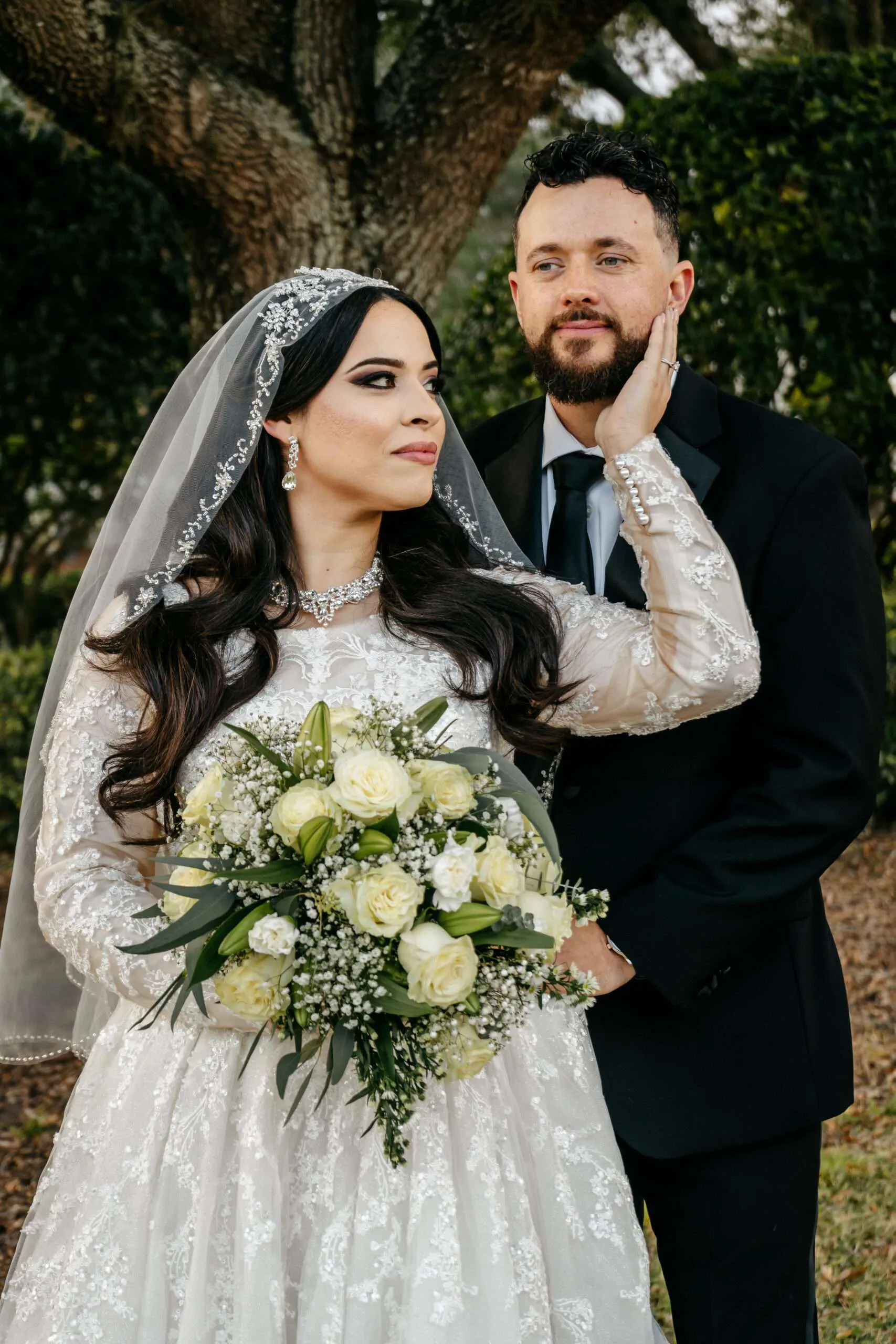 Bride in a beaded veil touching her groom's cheek beneath a sprawling tree