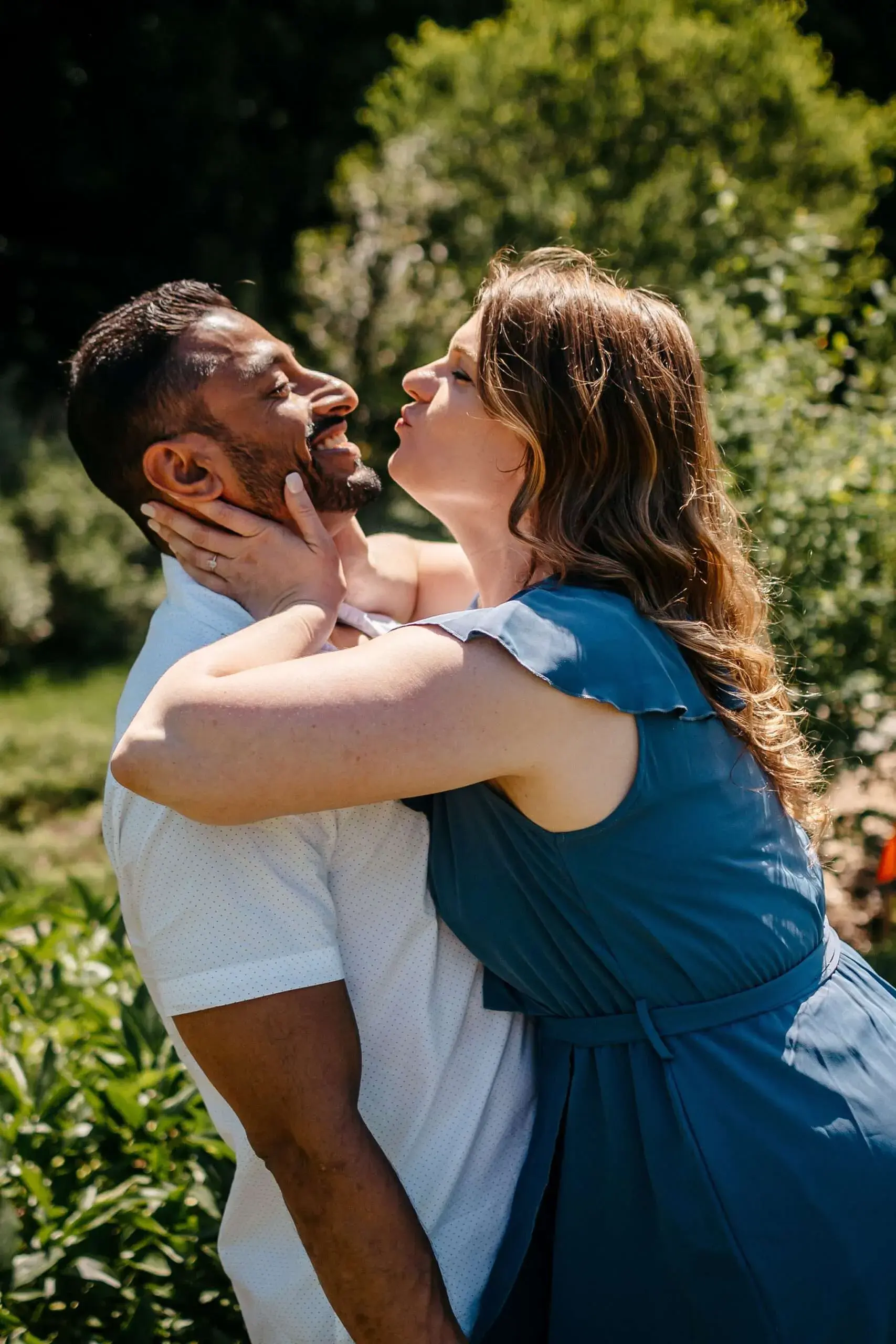 Intimate portrait of a woman kissing a man's cheek while embracing him in the sunny gardens of the Arnold Arboretum in Boston, MA.