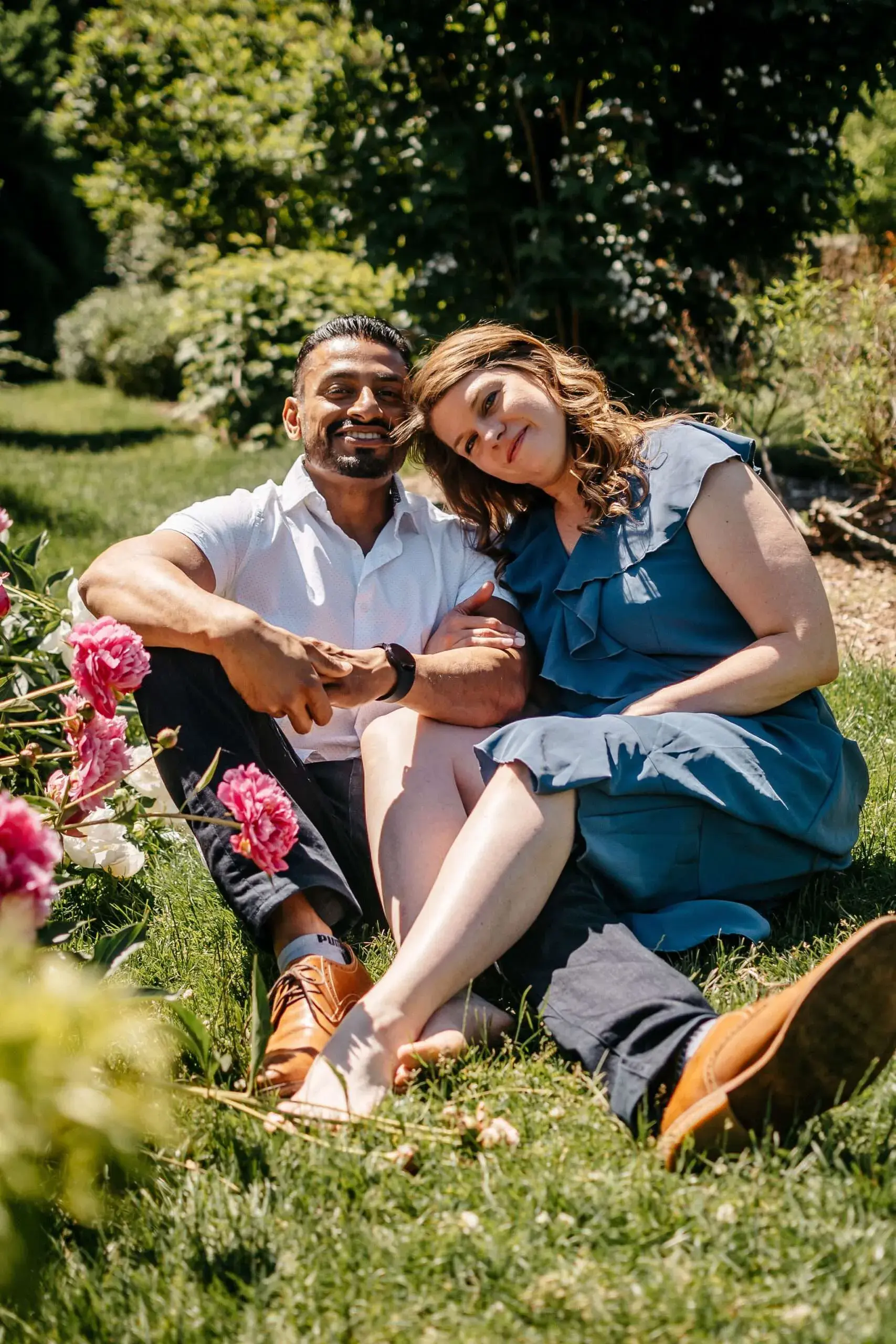 A relaxed portrait of a couple sitting on the grass near pink peonies on a sunny day at the Arnold Arboretum in Boston, MA.