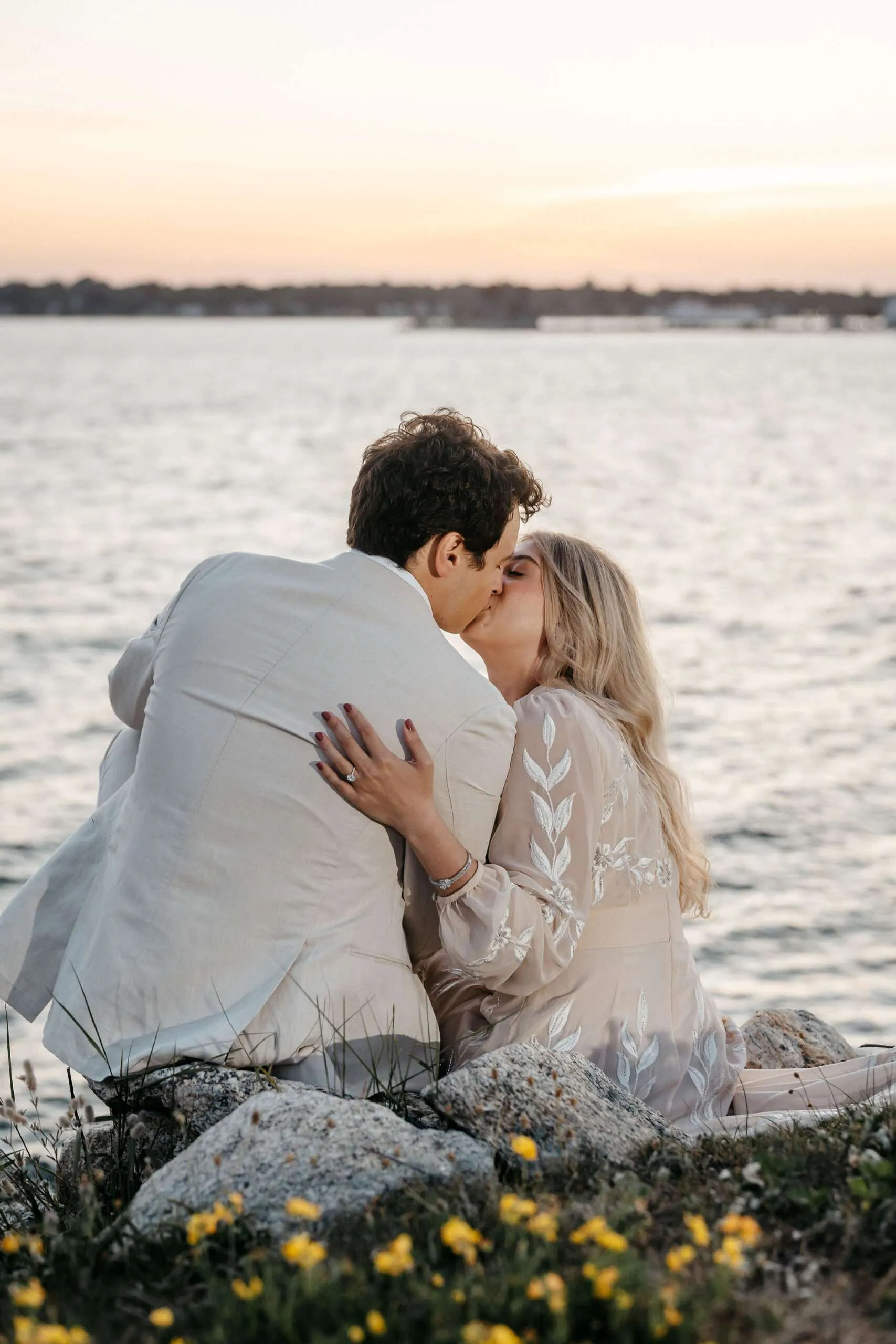 Patrick and Soumaya kissing at sunset by the water on a rocky shoreline surrounded by yellow wildflowers.