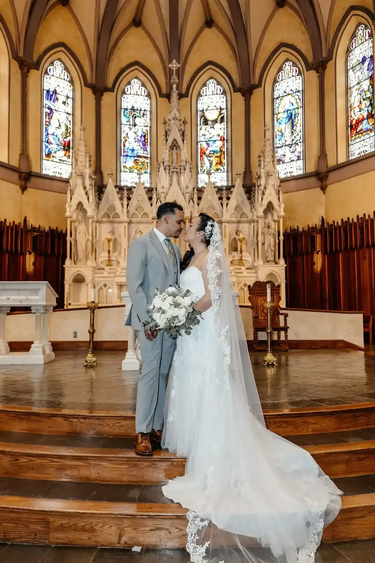 Bride and groom standing on the altar steps with stained glass windows in the background at Saint Patrick Parish in Lawrence, MA.