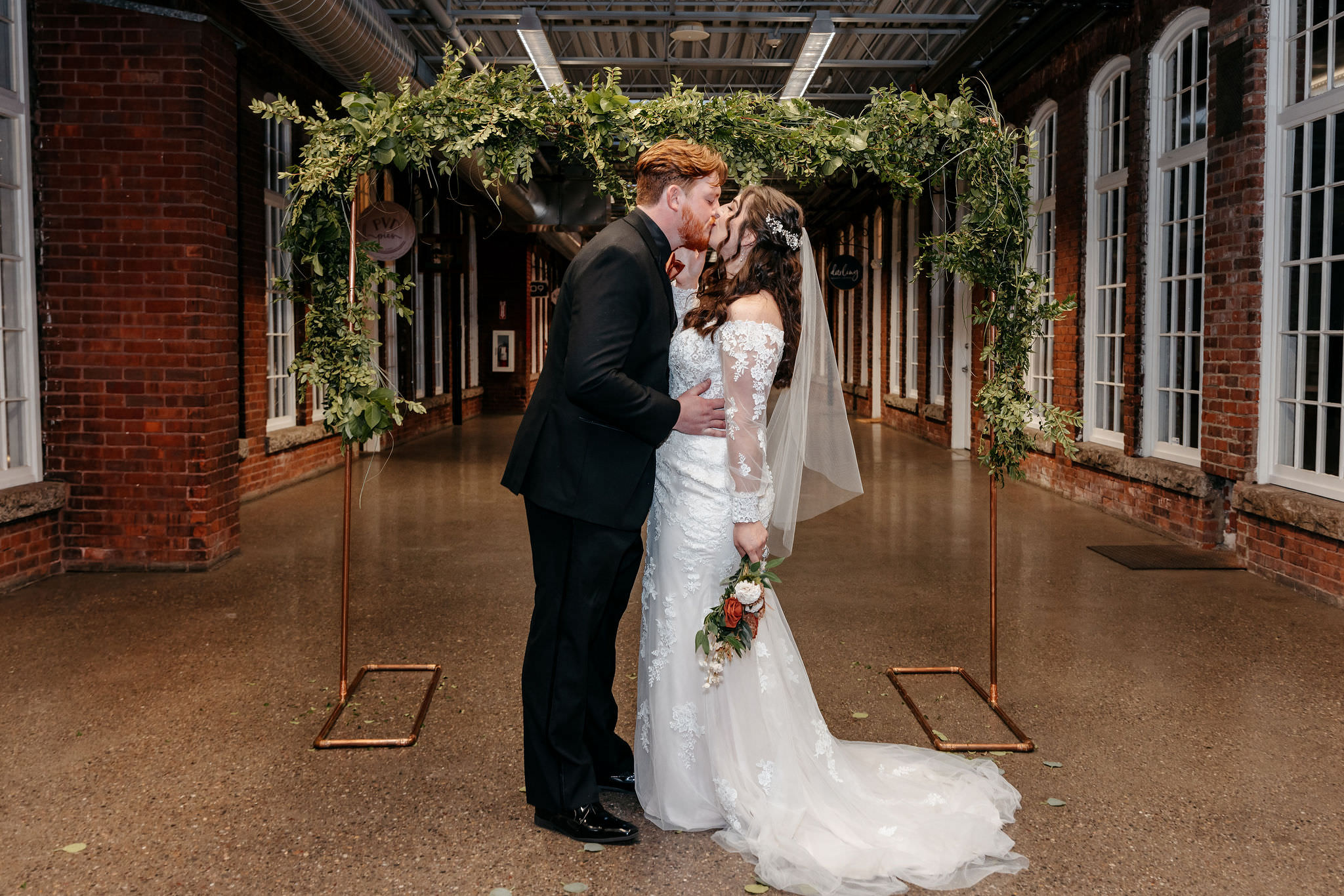 Couple kissing beneath a greenery arch in a brick industrial hall