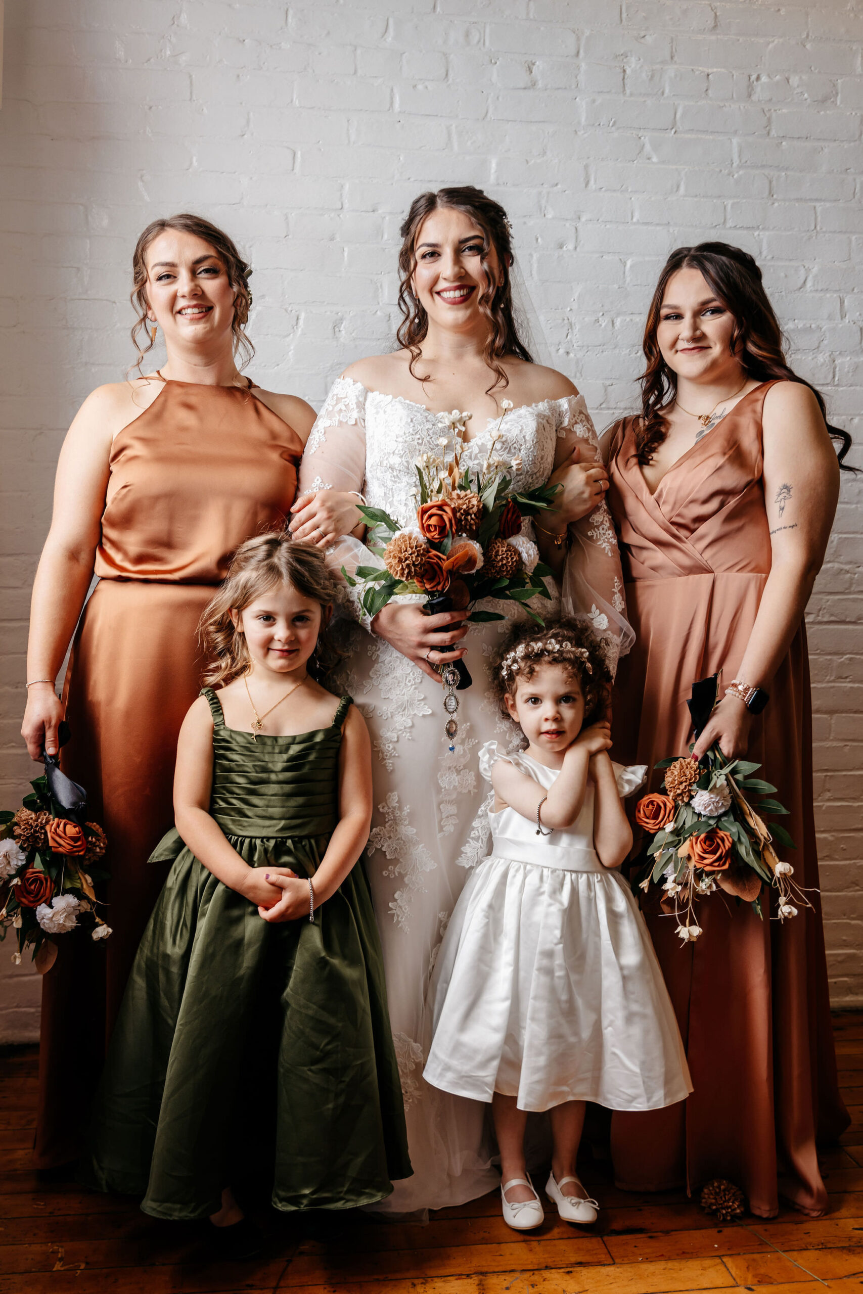 Bride with bridesmaids in copper dresses and flower girls against a white brick wall