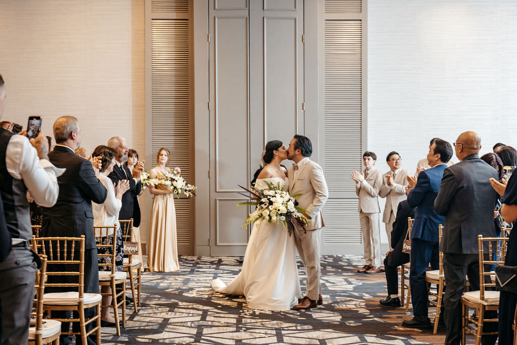 Bride and groom sharing their first kiss as guests applaud in the ballroom