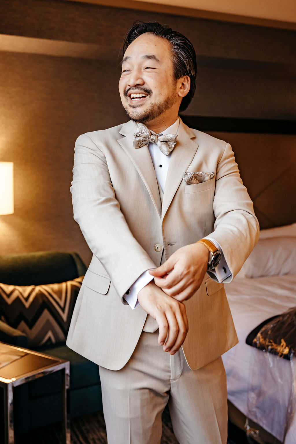 Groom in a tan suit and floral bow tie fastening his cufflinks while getting ready