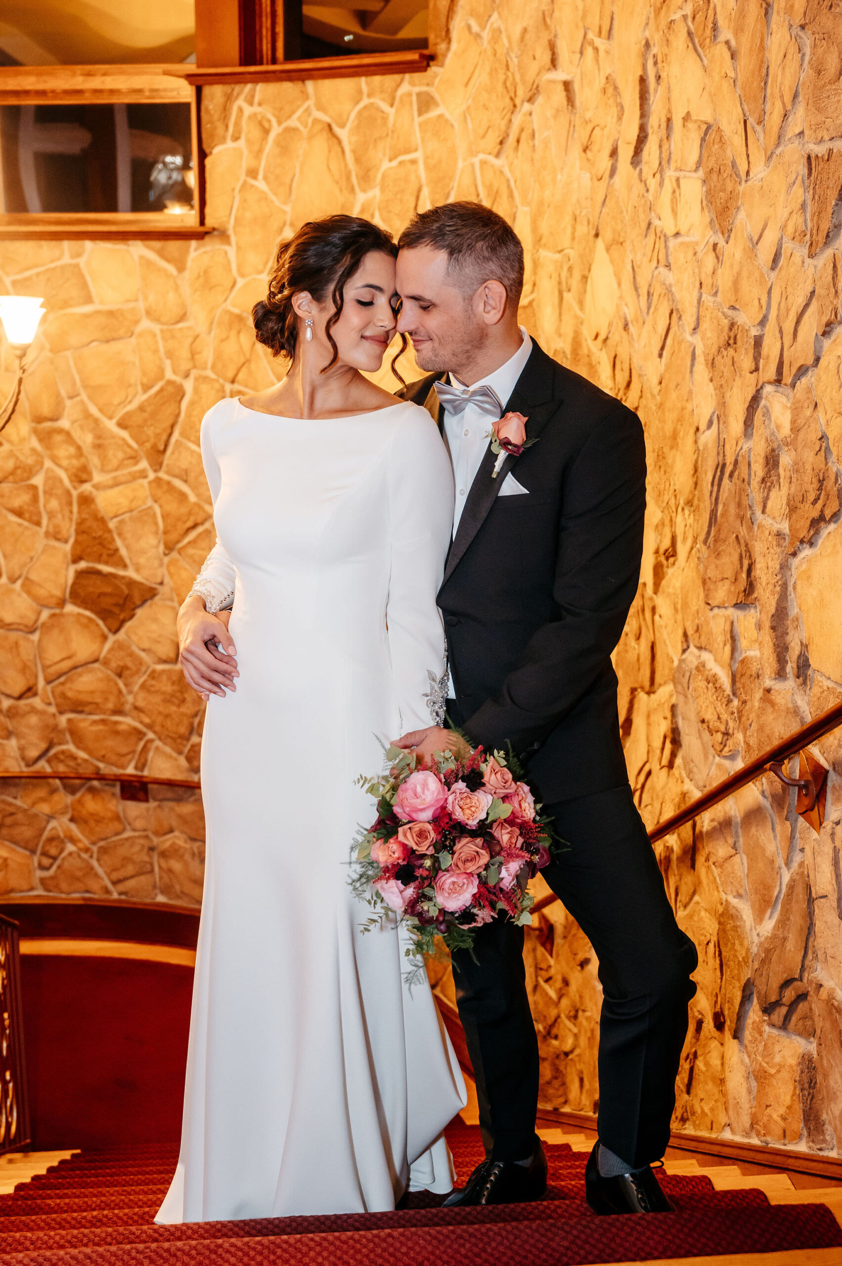 Bride and groom nuzzling on a staircase with a pink rose bouquet