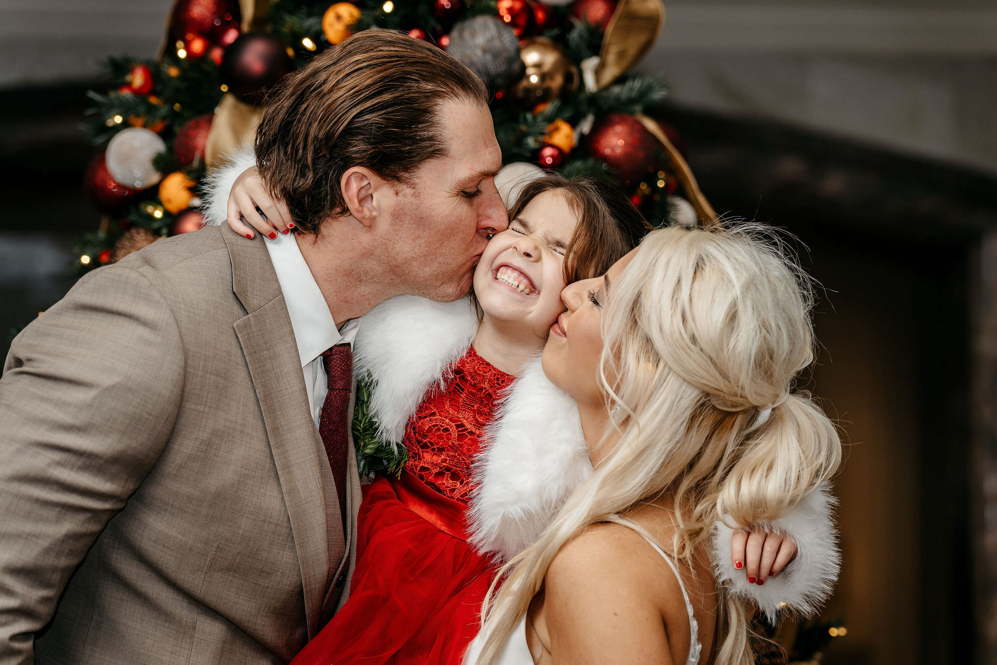 Parents kissing their daughter's cheeks in front of a Christmas tree
