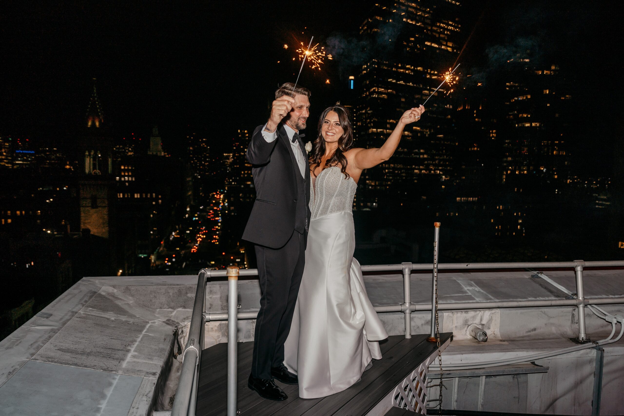 Bride and groom holding sparklers on rooftop overlooking city skyline at Boston hotel wedding venue at night