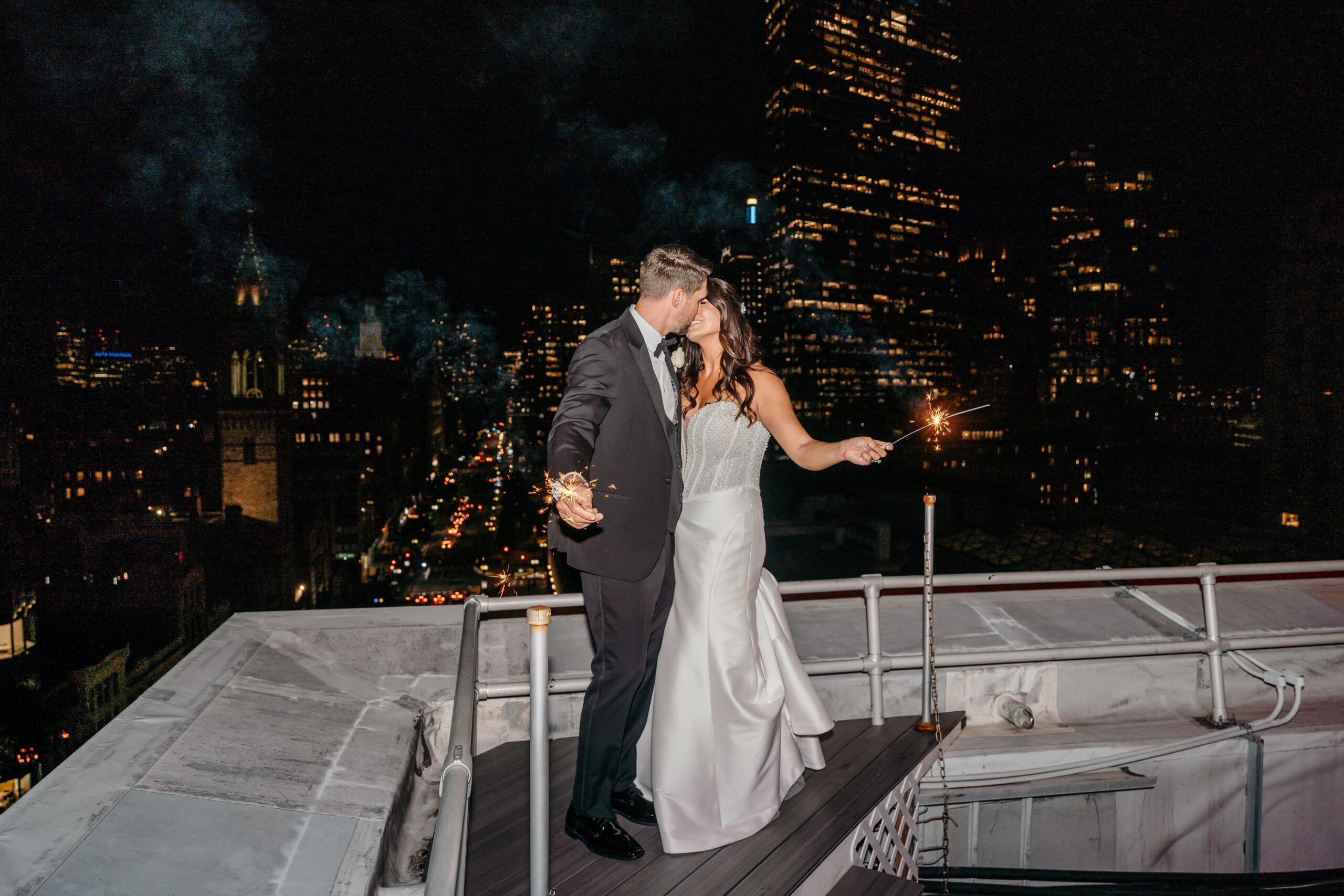 Bride and groom kissing with sparklers on rooftop at Boston hotel wedding venue with nighttime city views