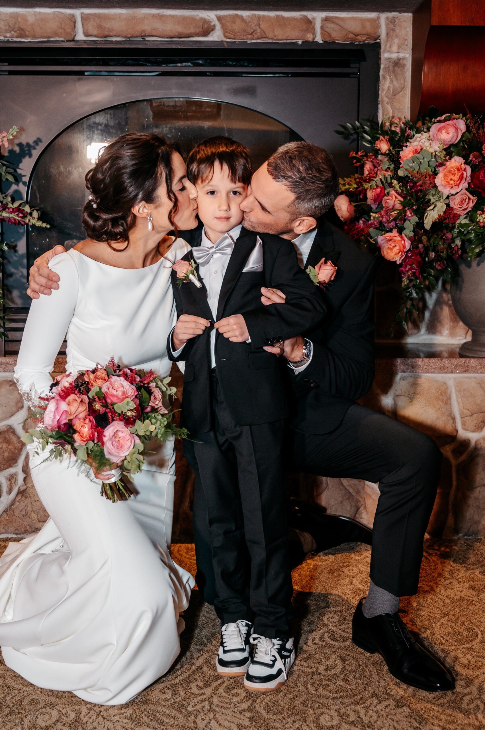 Bride and groom kissing child on cheeks during Burlington wedding at Cafe Escadrille, candid family moment with floral backdrop
