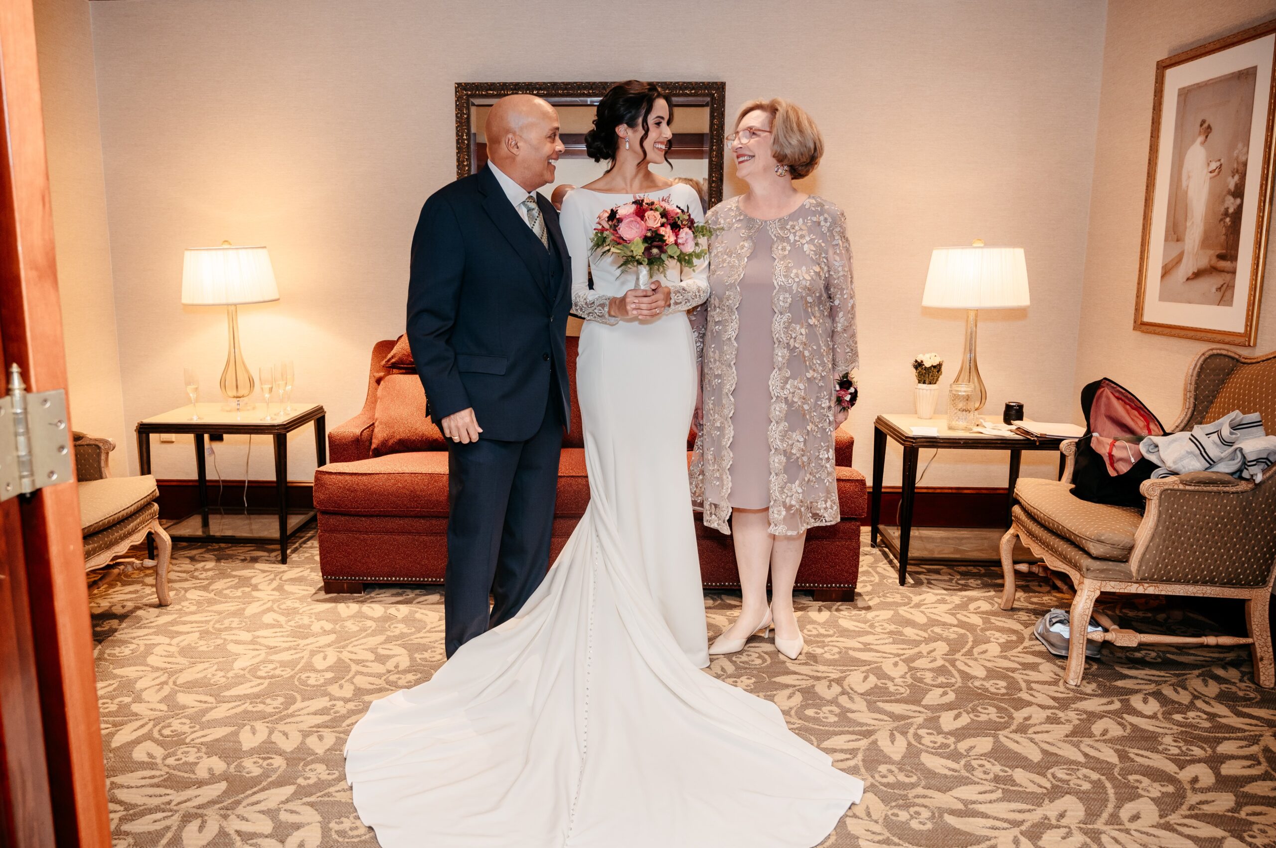 Bride smiling with parents in getting-ready suite before Burlington wedding at Cafe Escadrille, full-length gown and bouquet visible