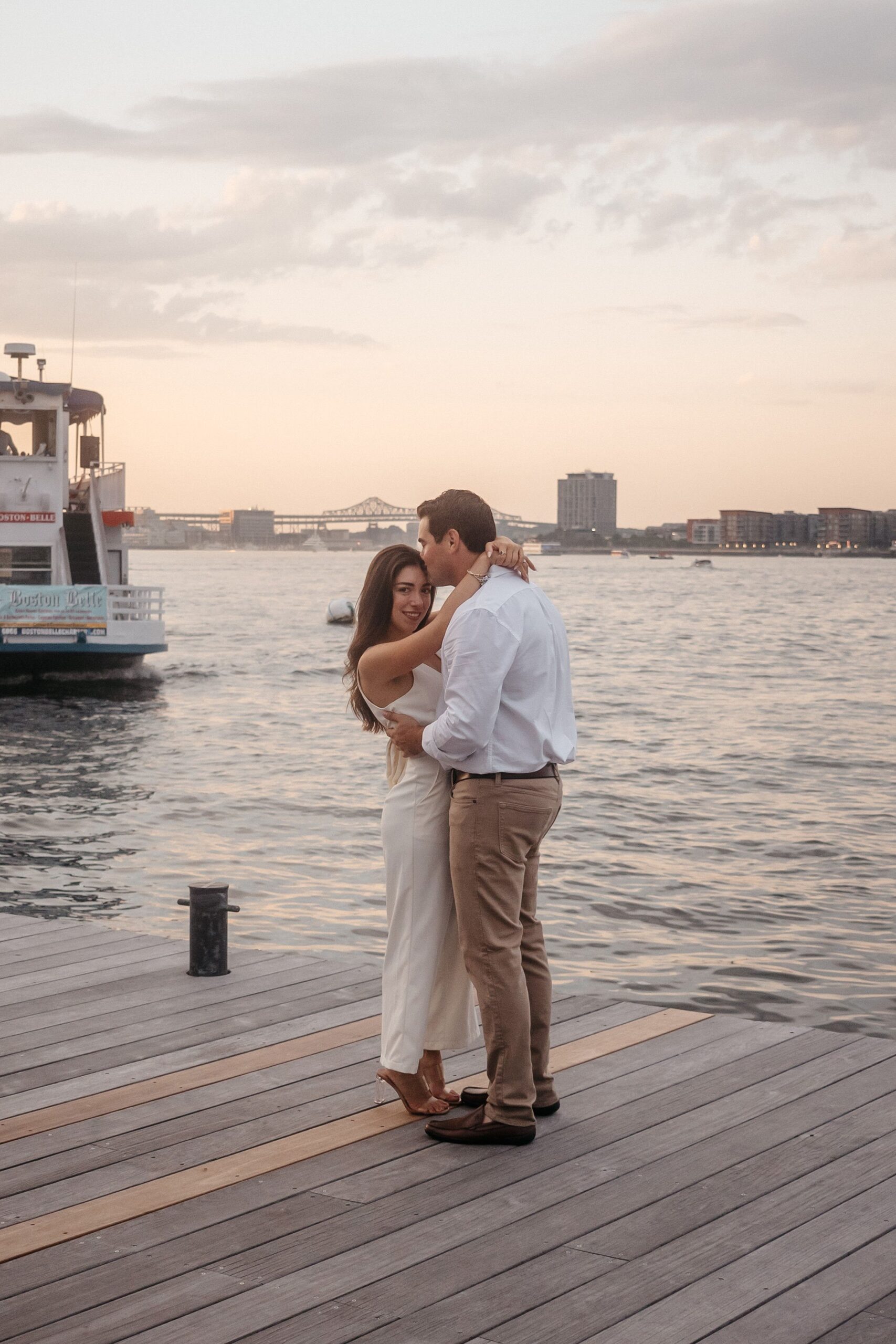 Couple embracing at Fan Pier Park Boston during engagement session, scenic harbor view with sunset and city skyline