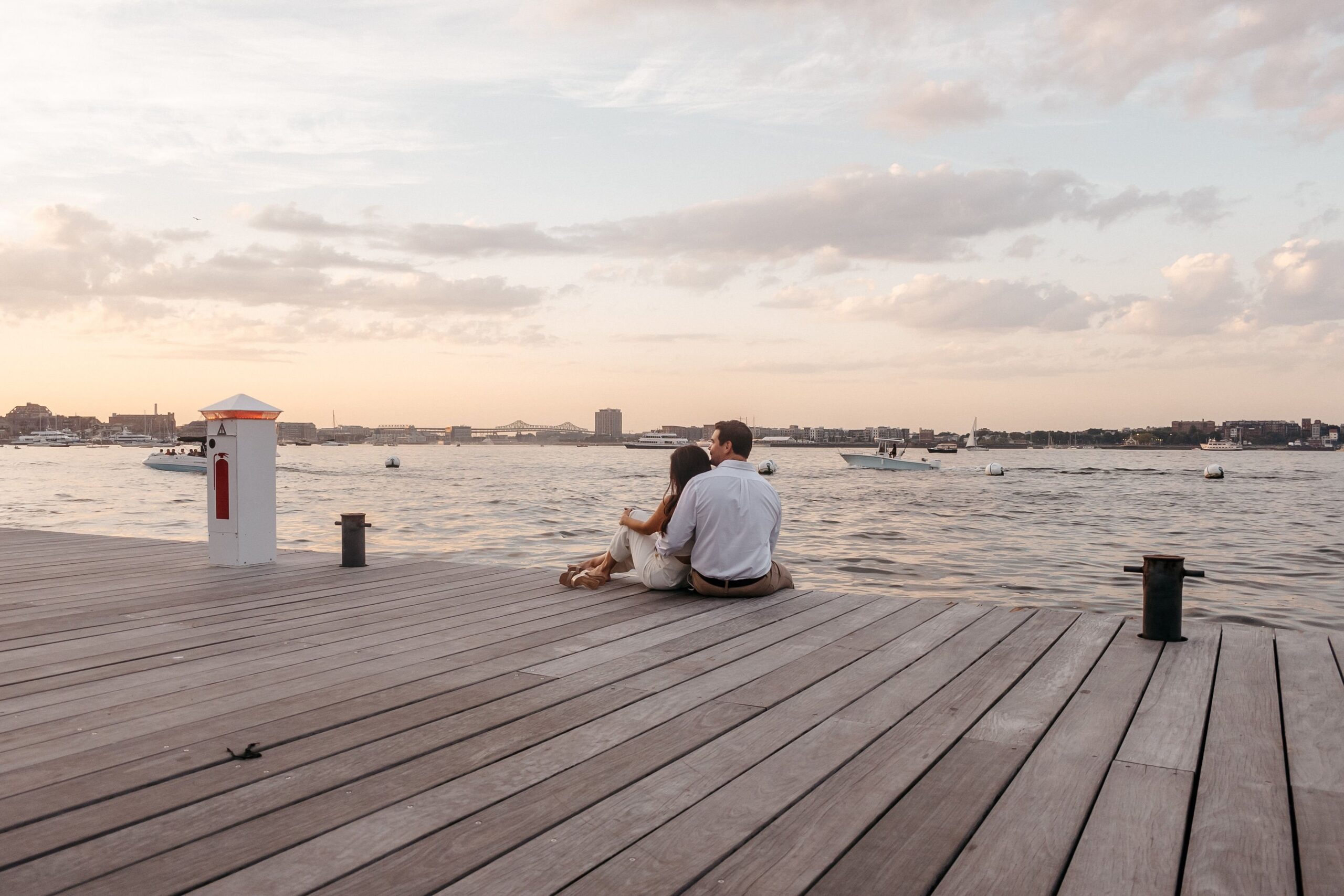 Couple sitting on the dock at Fan Pier Park Boston during engagement session, wide waterfront view at sunset