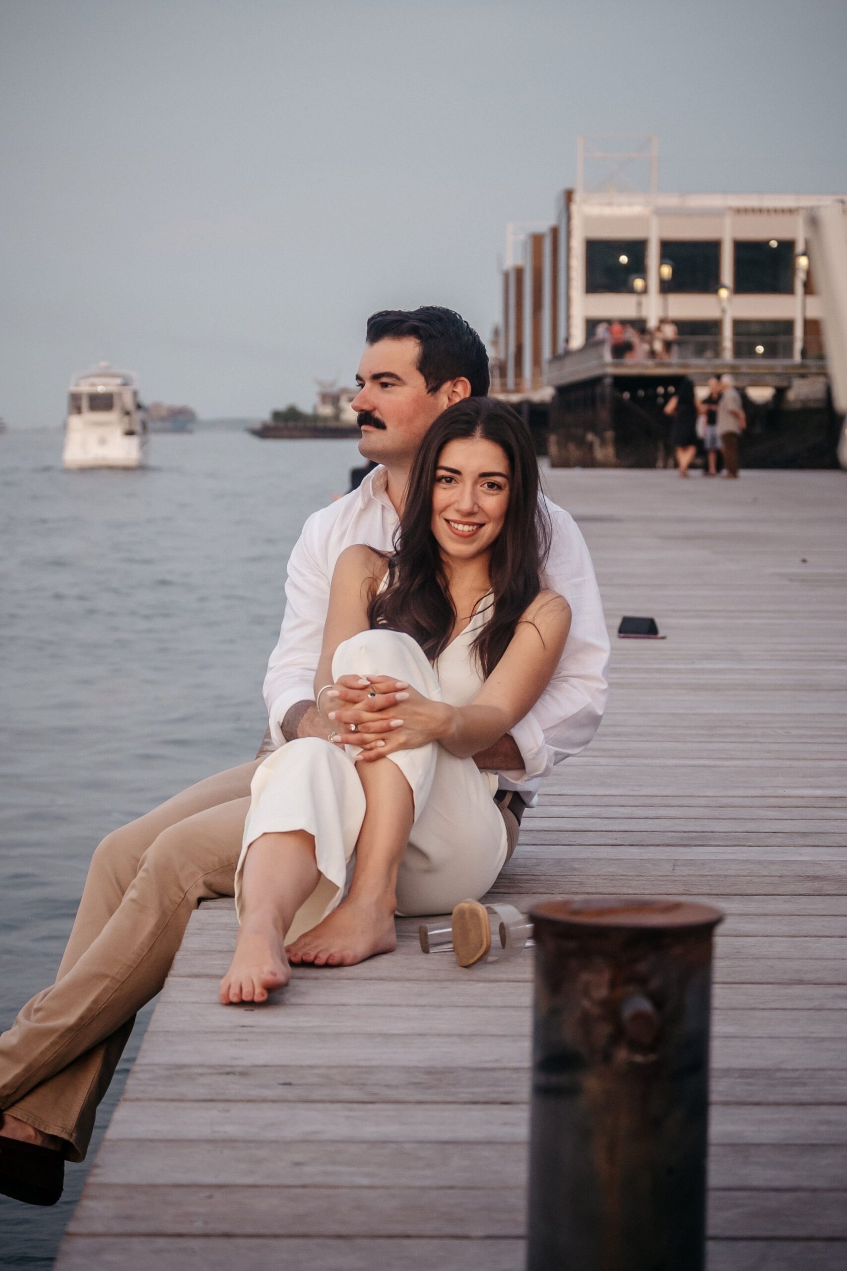 Couple seated along the harbor at Fan Pier Park Boston engagement session, soft evening light over the water