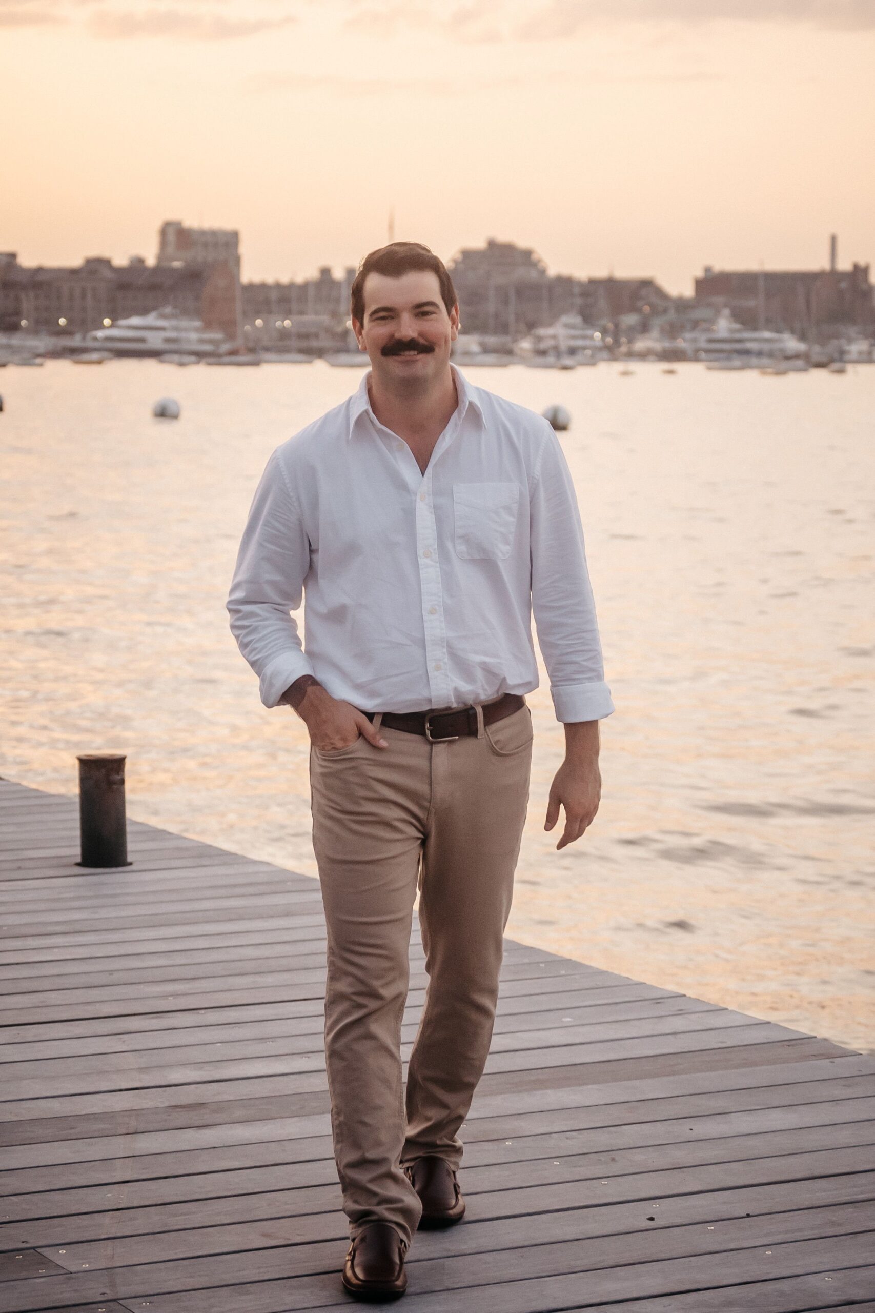 Groom walking along Fan Pier Park pier during engagement session, relaxed Boston waterfront at sunset