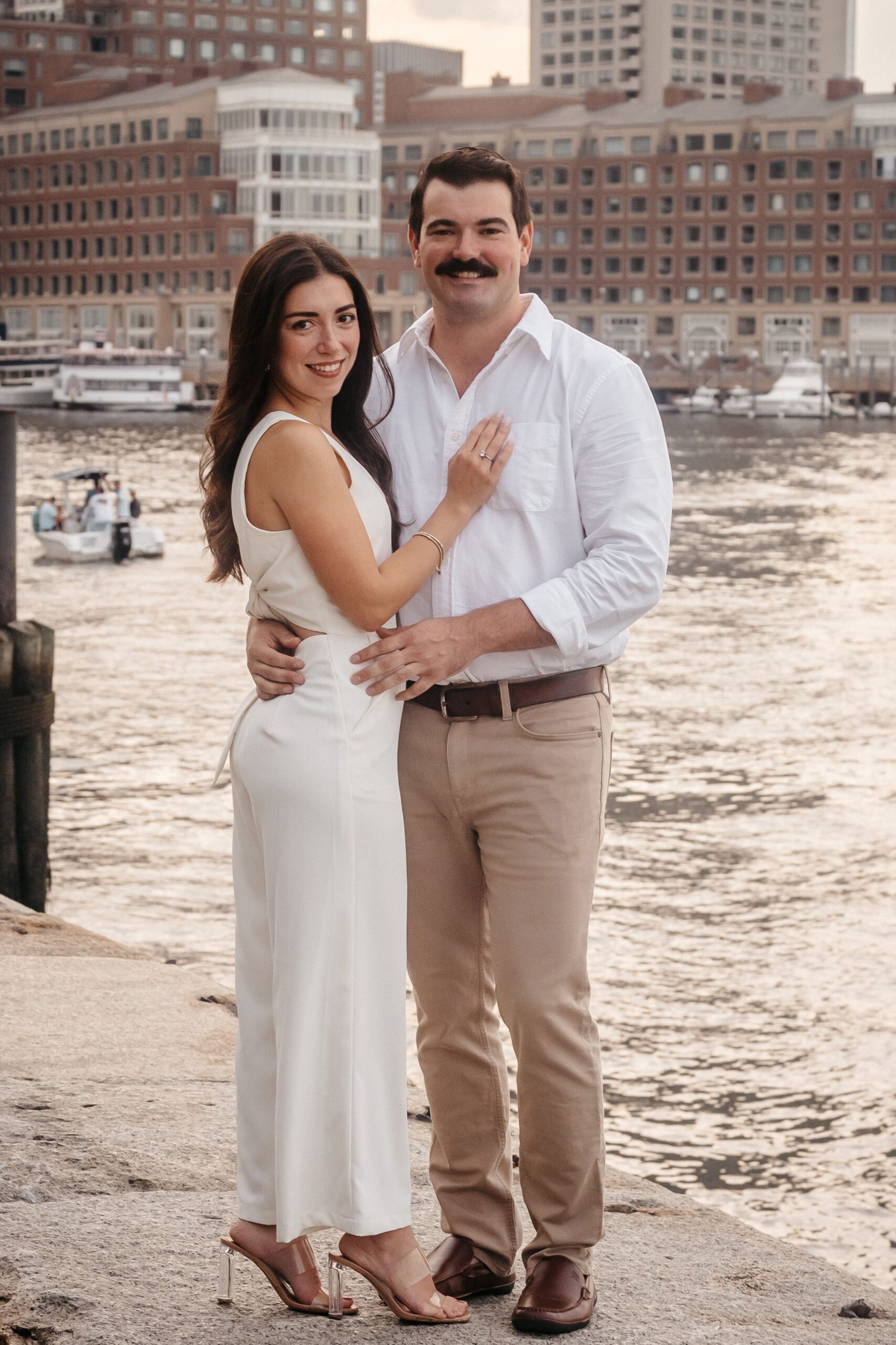 Engaged couple posing along the waterfront at Fan Pier Park Boston, classic engagement session portrait with city skyline backdrop