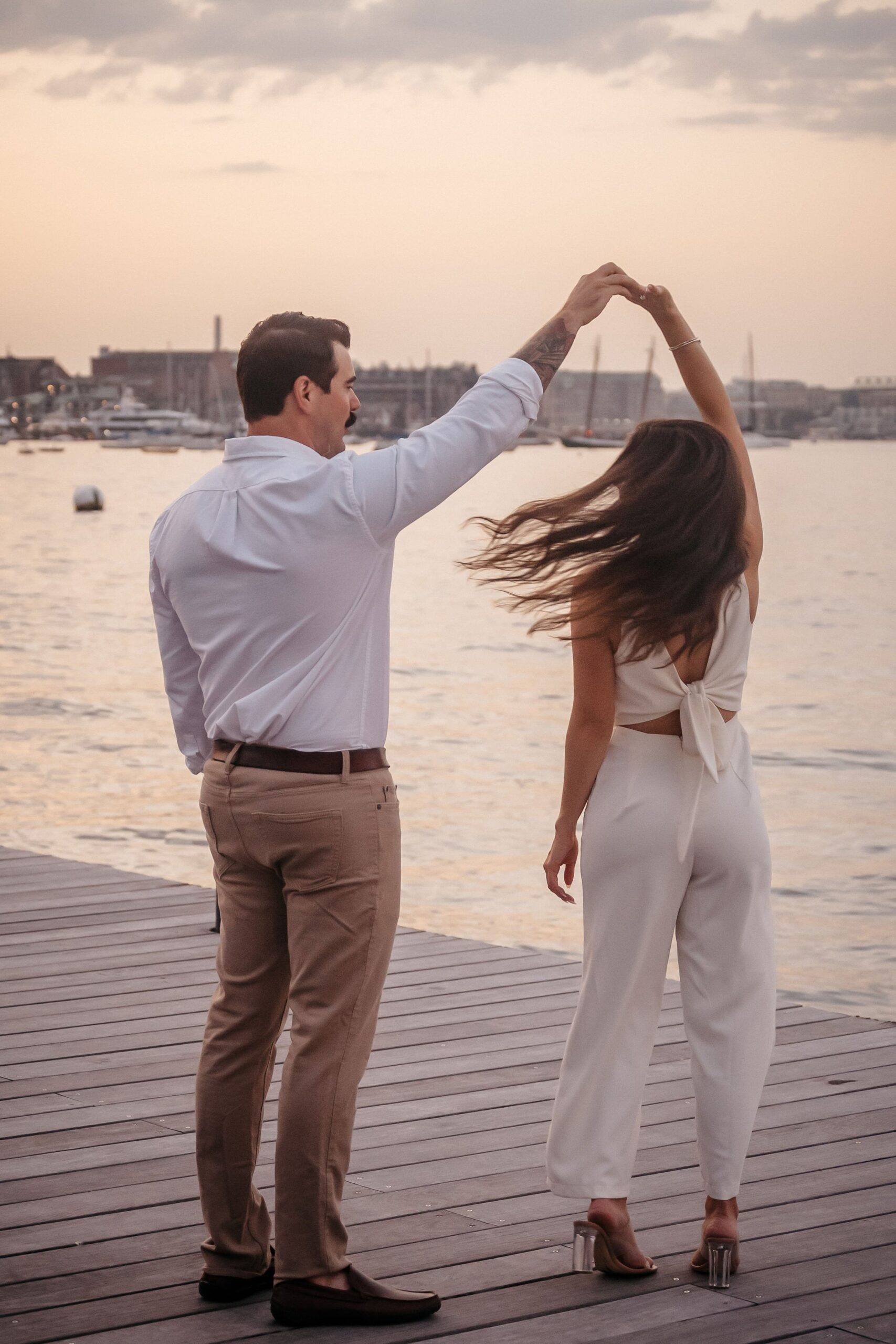 Couple twirling on the pier during Fan Pier Park engagement session, playful waterfront moment at sunset in Boston