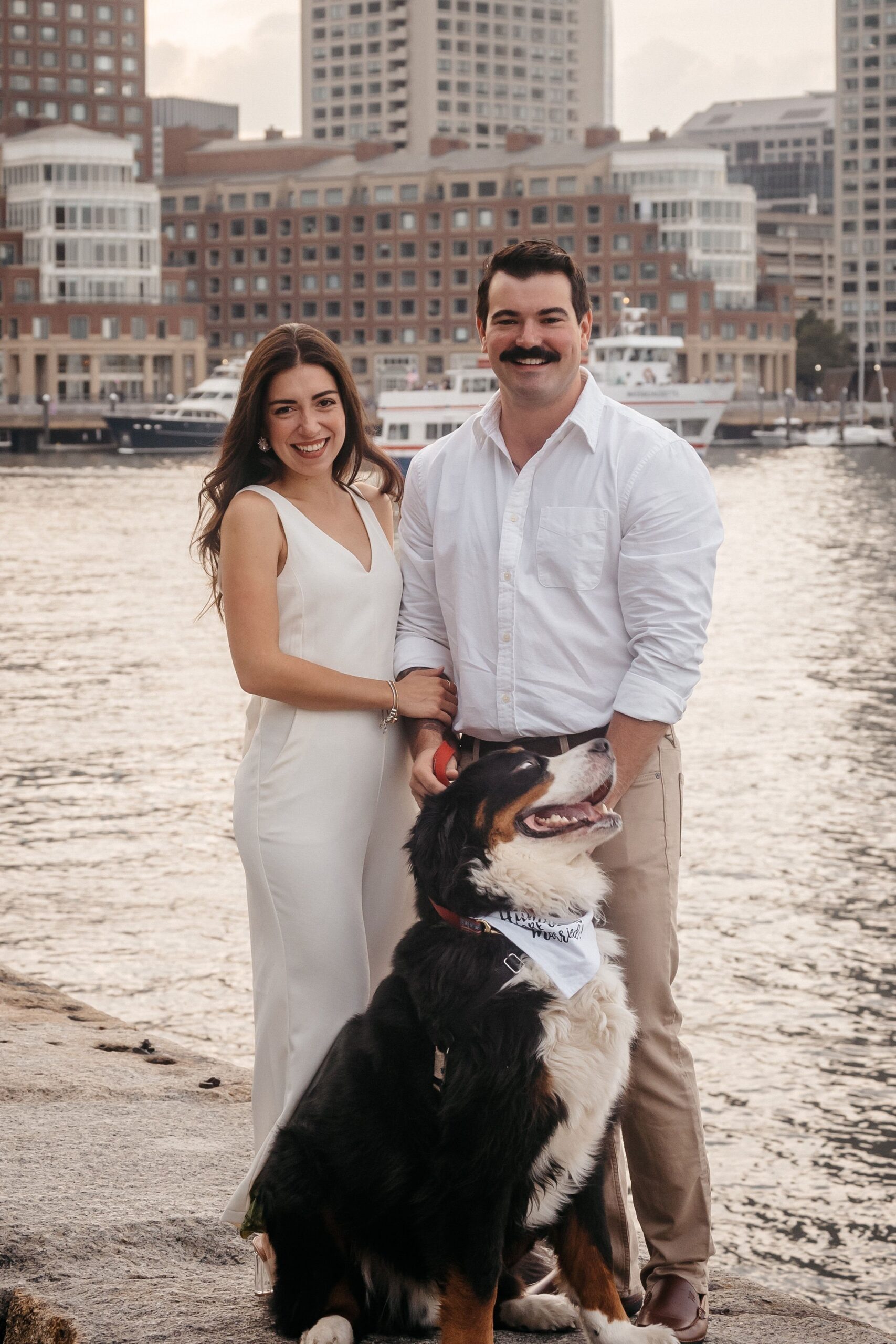 Couple with their dog at Fan Pier Park Boston engagement session, waterfront skyline backdrop during soft sunset light