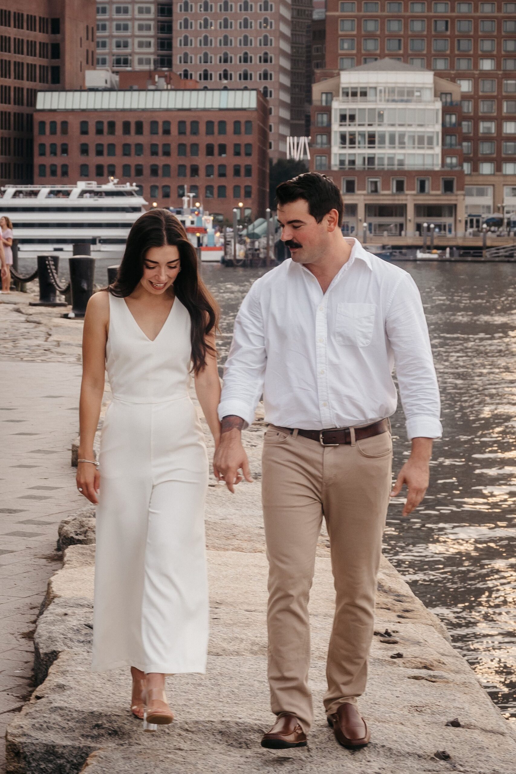 Couple walking along the waterfront at Fan Pier Park Boston during engagement session, candid moment by the harbor